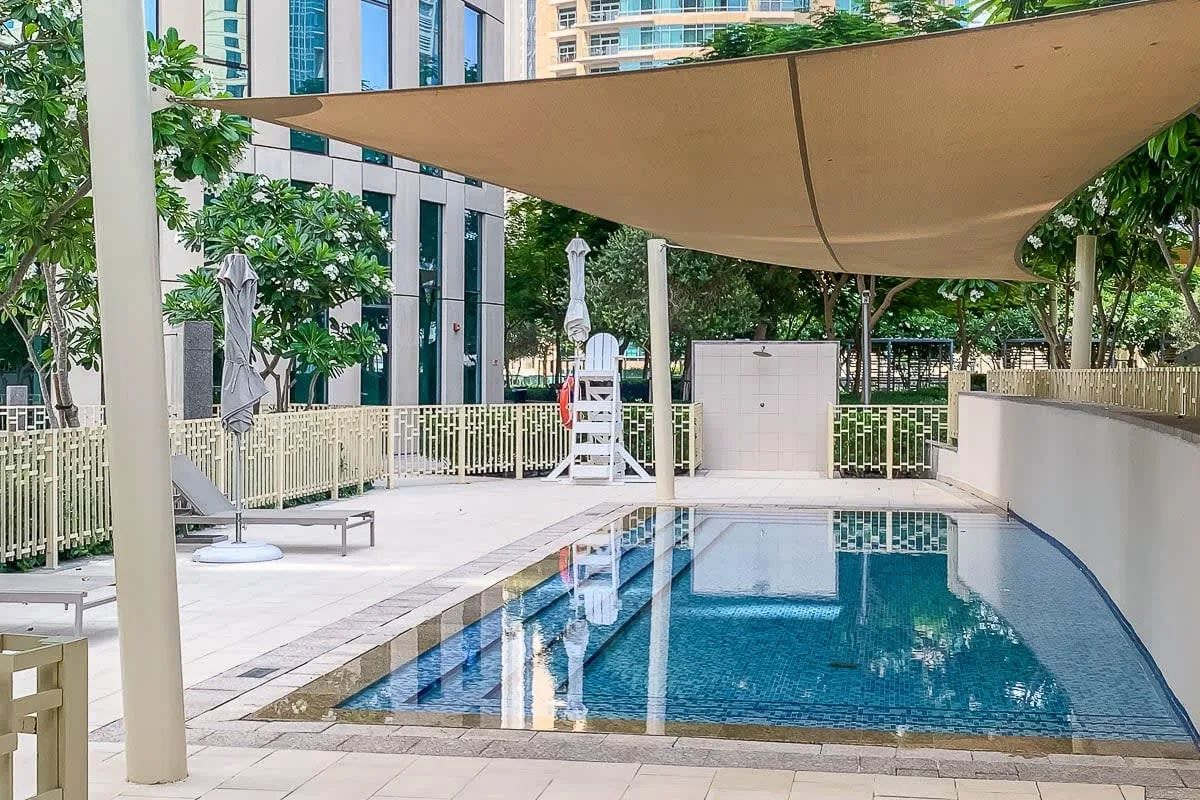 An outdoor swimming pool sits beneath a large shade canopy, with trees and buildings nearby.