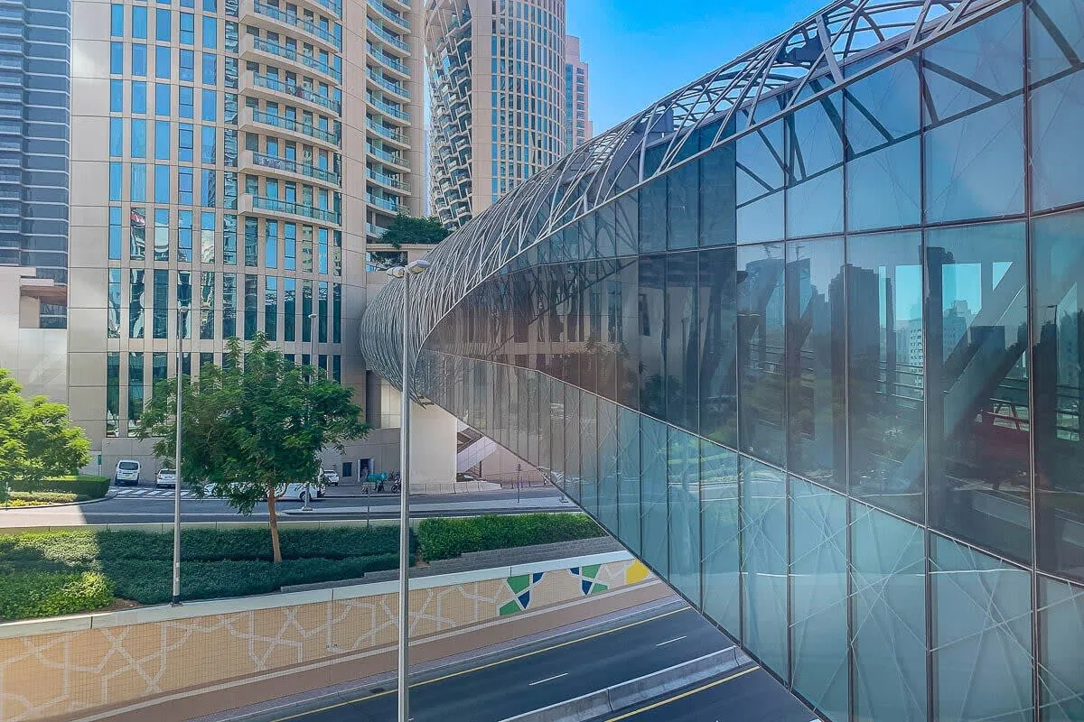 A glass pedestrian bridge curves between modern office buildings above a landscaped urban plaza.