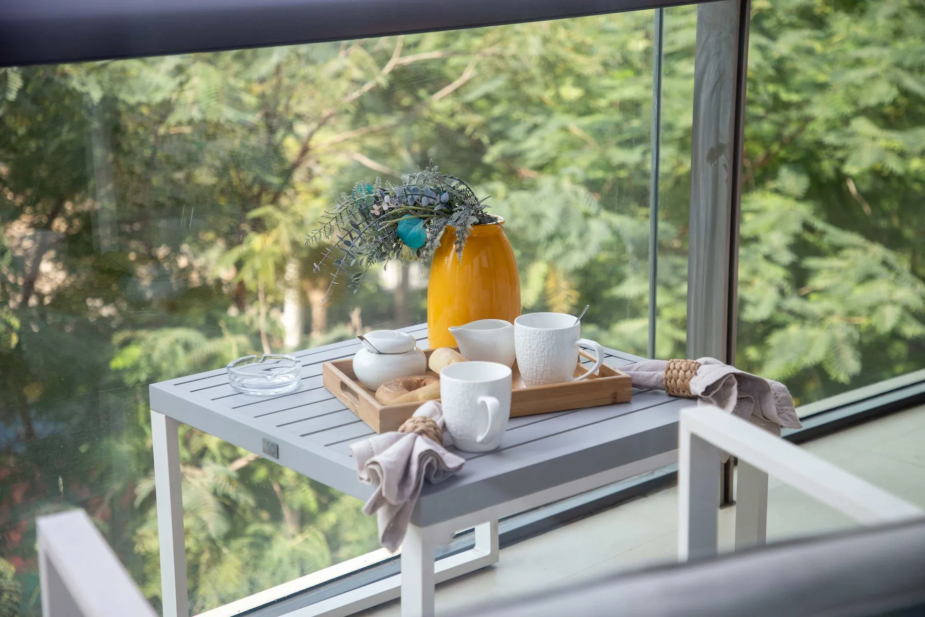 A tea setting rests on a table beside a large window overlooking leafy trees.