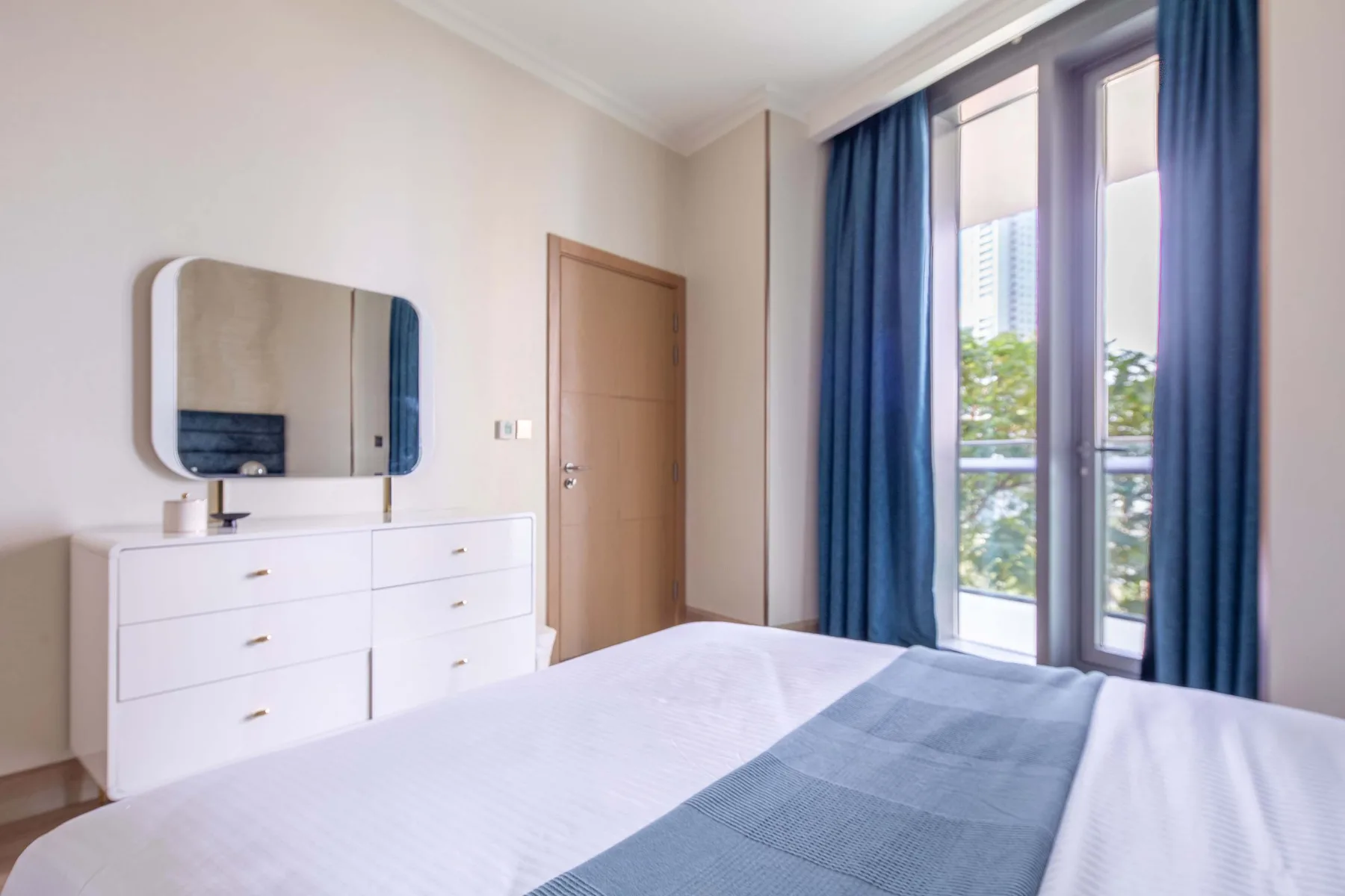 A bedroom with a white bed, dresser, and tall windows with blue curtains.