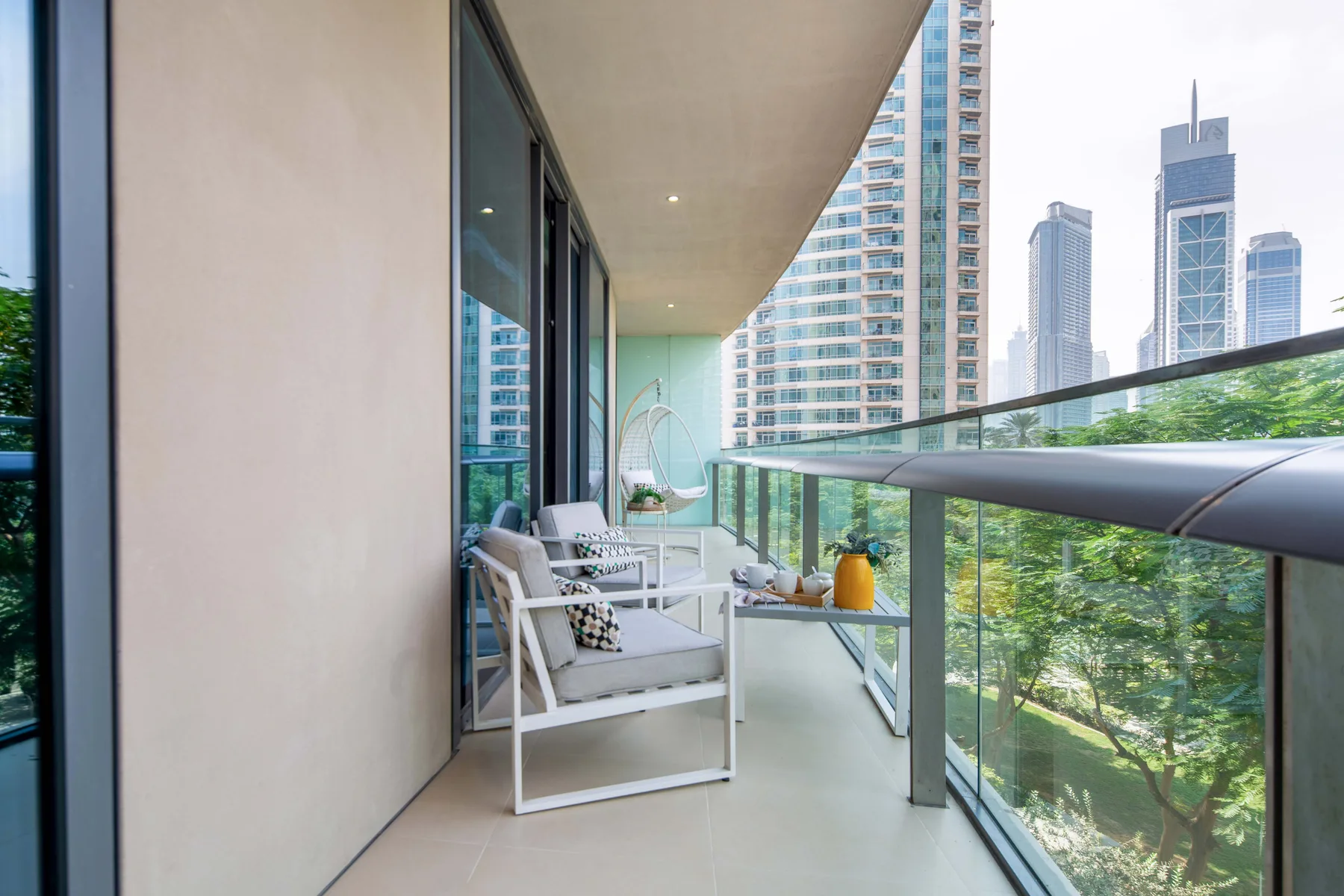 A balcony with outdoor seating overlooks trees and nearby high-rise buildings.