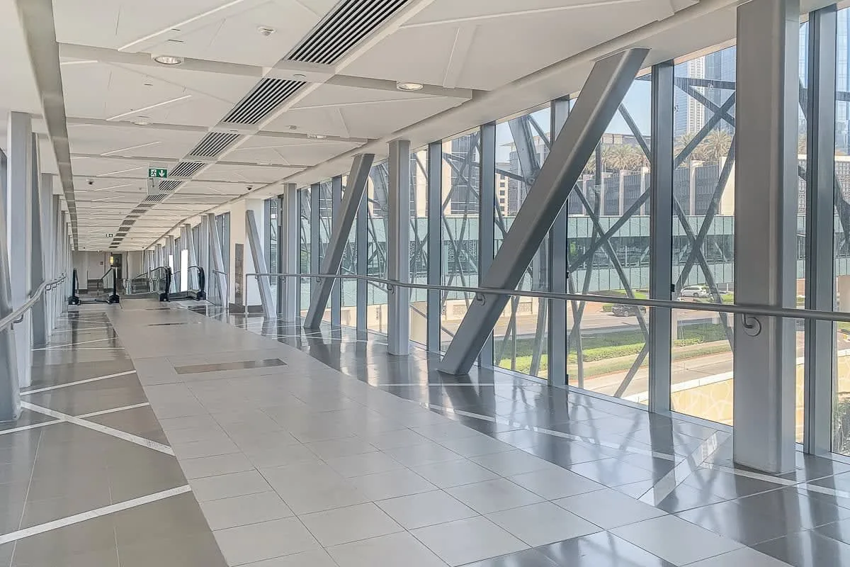 A bright indoor walkway with large windows and structural beams along one side.
