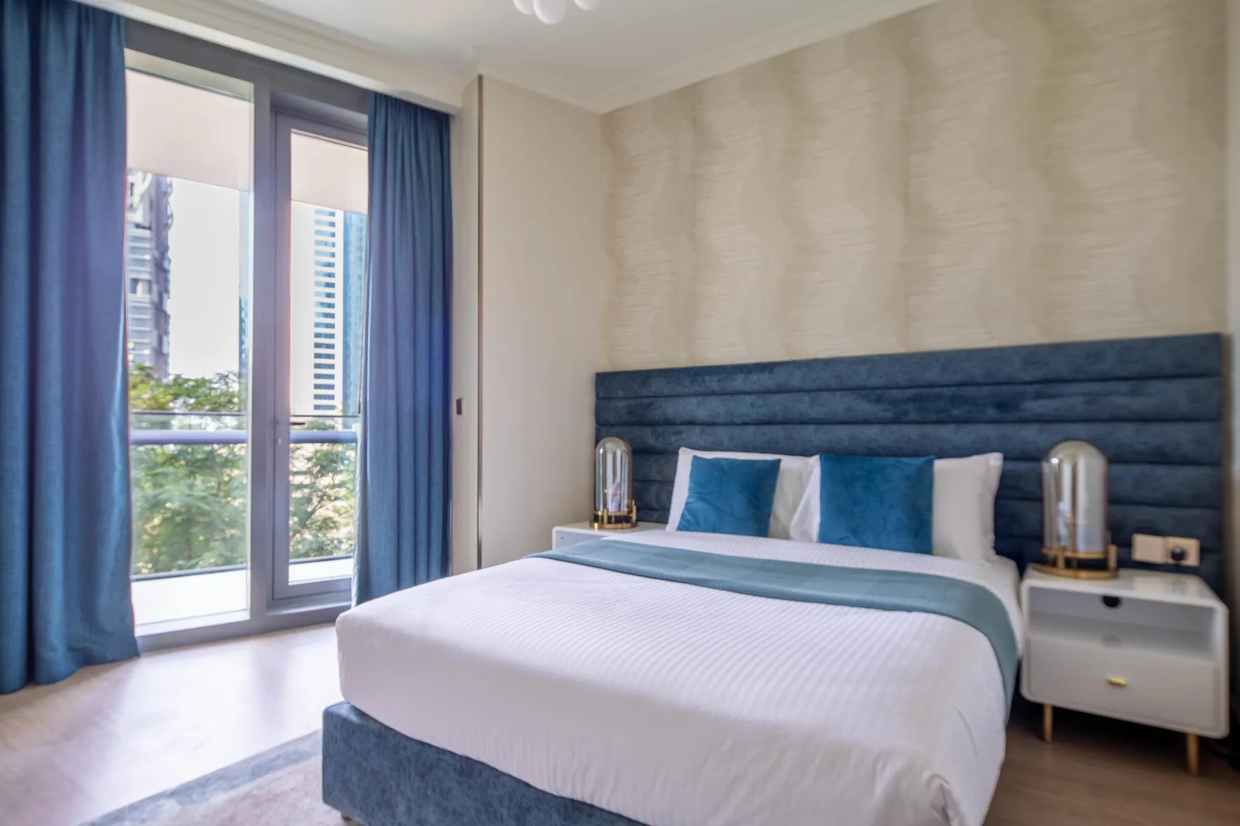 A bedroom with a neatly made bed and blue curtains beside tall windows.