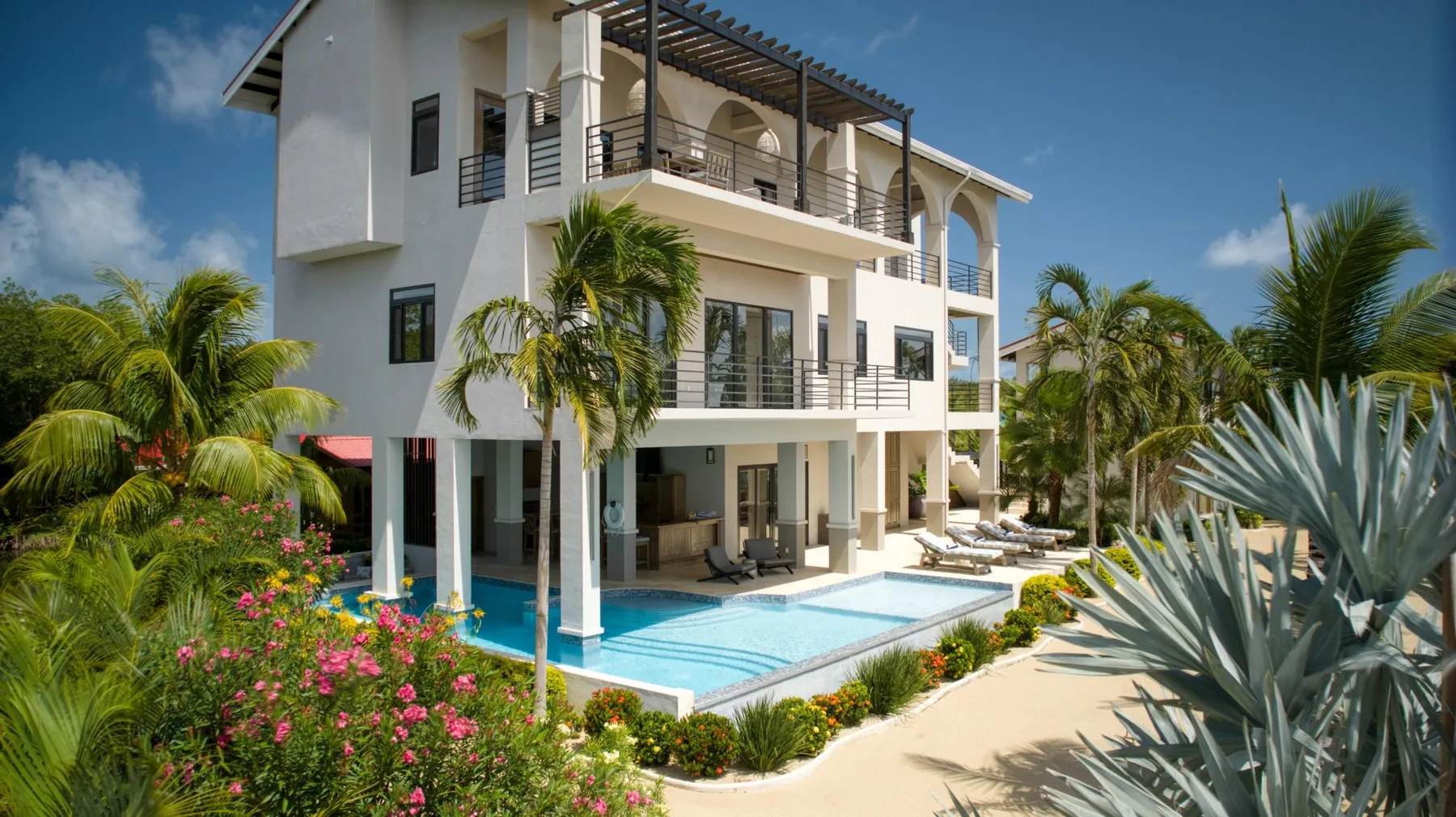 A modern white villa overlooks a swimming pool surrounded by tropical landscaping.