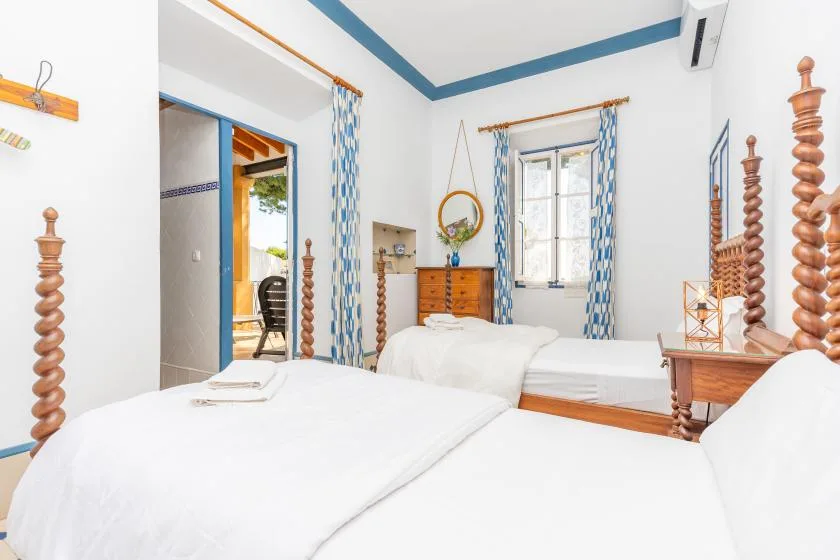 A bright bedroom with white bedding, blue trim, and a window overlooking greenery.