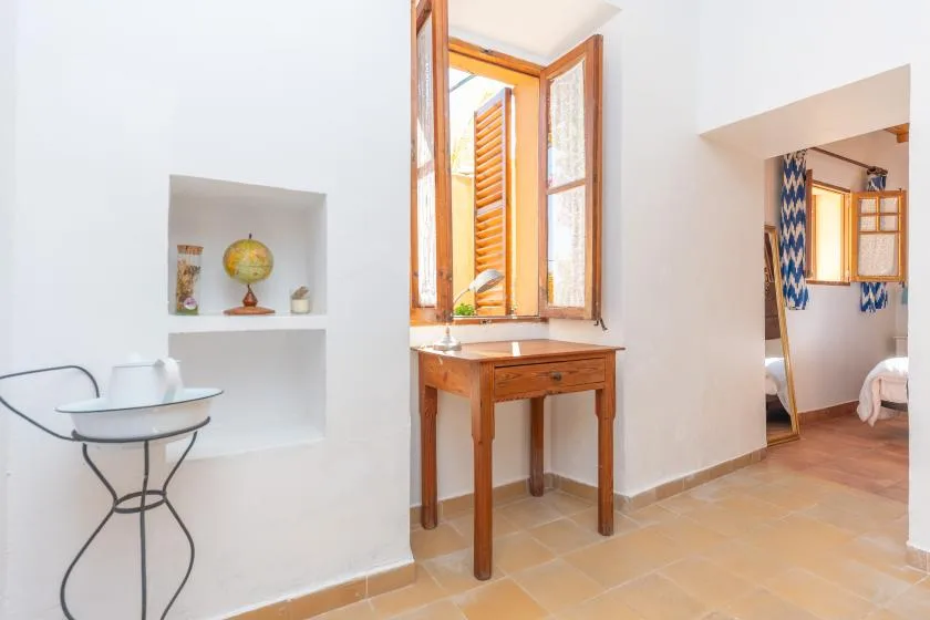 A bright interior with a wooden table beneath an open window and a doorway to a bedroom.