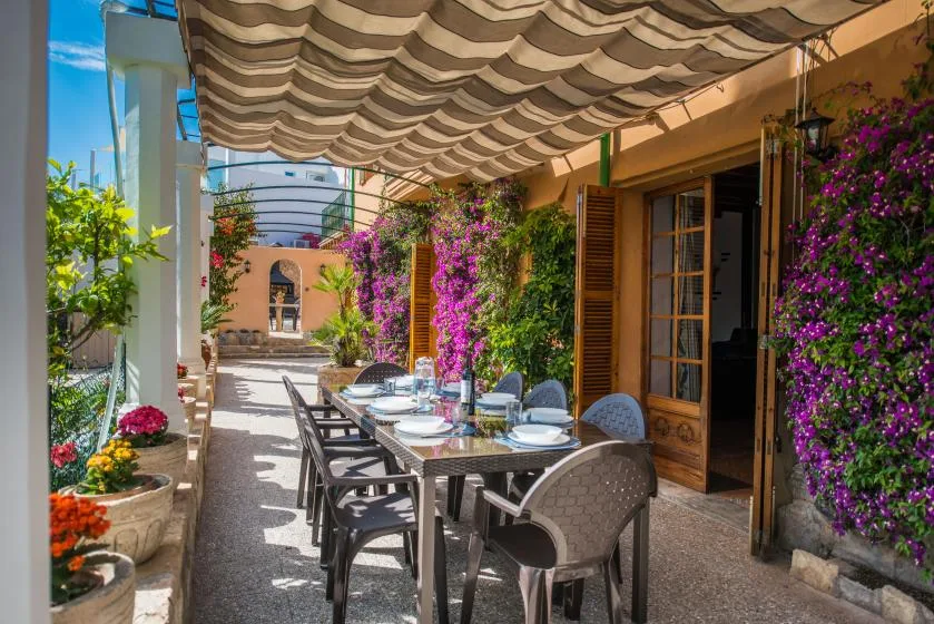 A shaded outdoor dining area with a long table beside flowering plants.