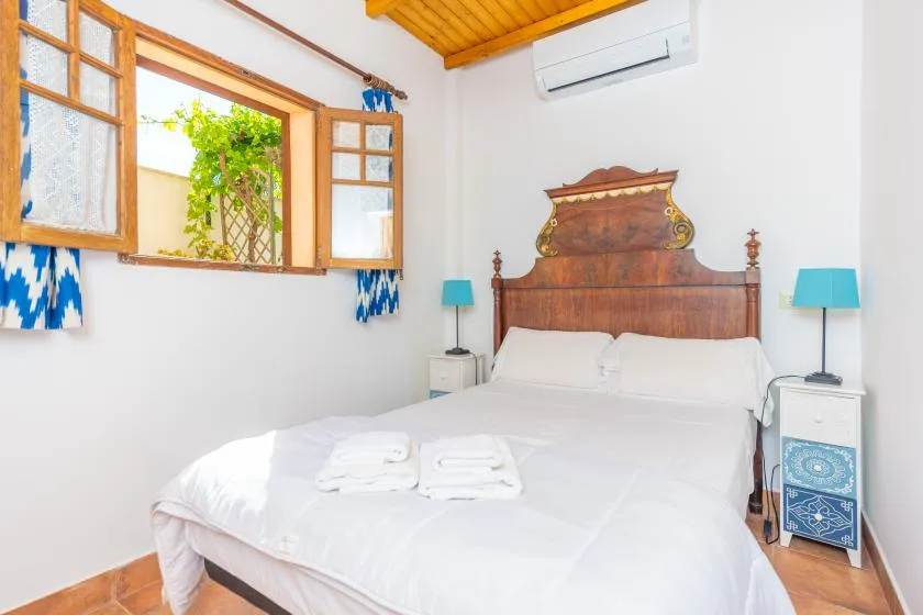 A bright bedroom with a double bed, wooden headboard, and open windows.