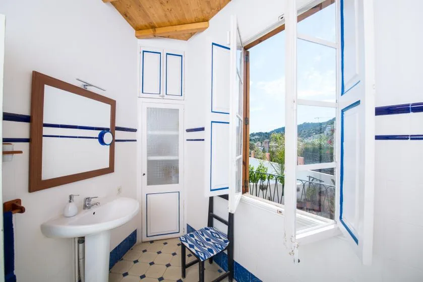 A bright bathroom with a sink beside open windows overlooking a green landscape.