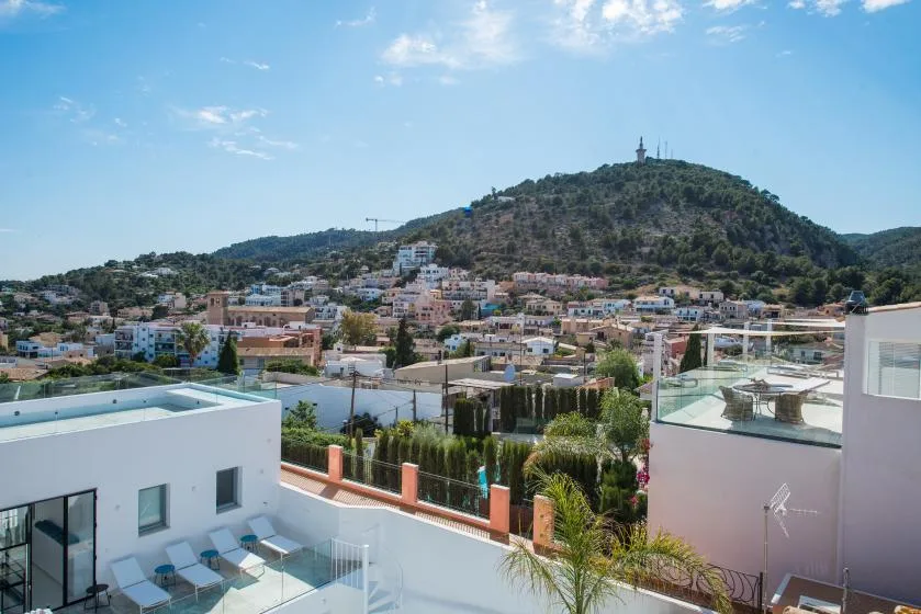 A hillside town rises behind modern rooftops and a rooftop pool under a blue sky.