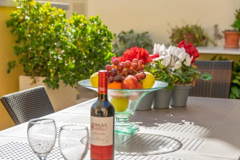 A patio dining table displays a fruit bowl, wine, and flowering plants.