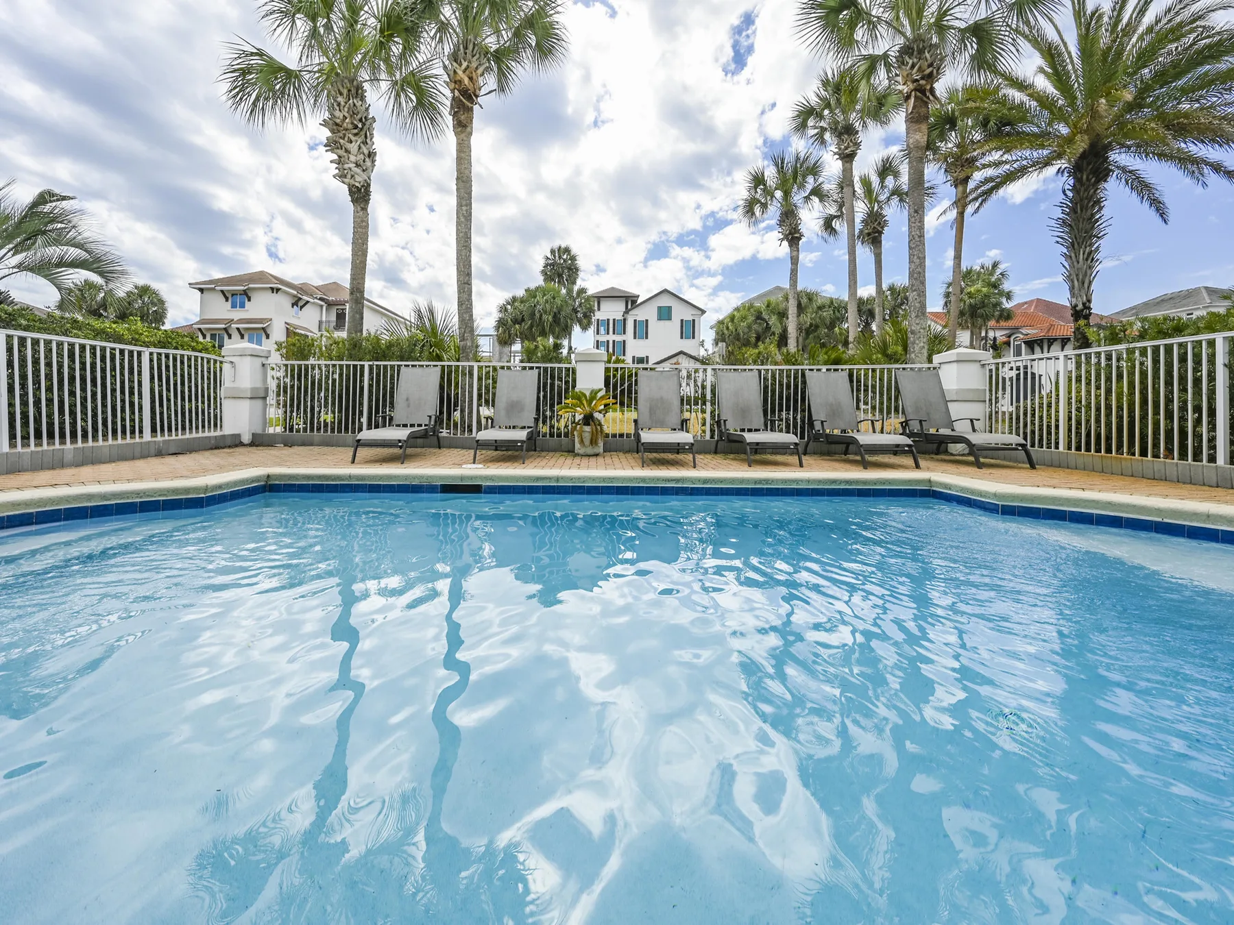 An outdoor swimming pool is bordered by lounge seating and palm trees.
