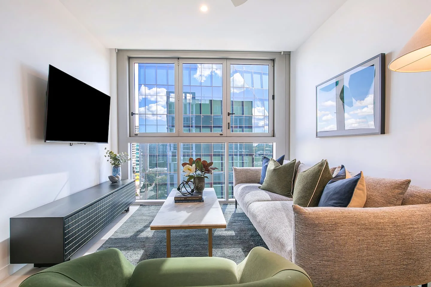 A bright living room with seating, a wall-mounted television, and large windows.