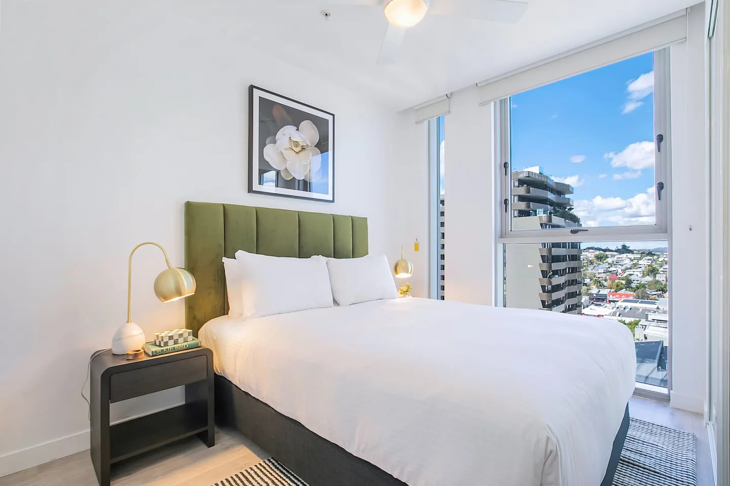 Un dormitorio luminoso con una cama blanca y grandes ventanas con vistas a edificios cercanos.
