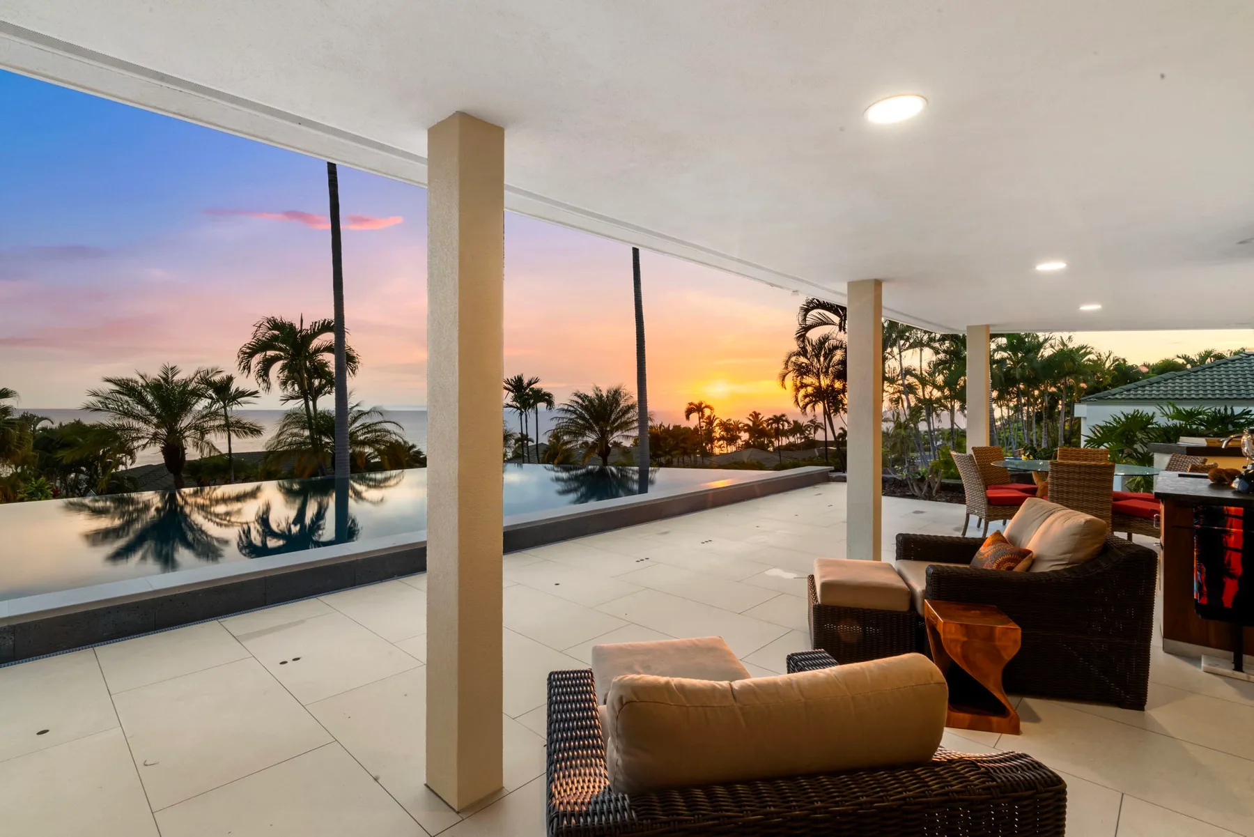 Una terraza cubierta da a una piscina y palmeras al atardecer, con asientos cerca.
