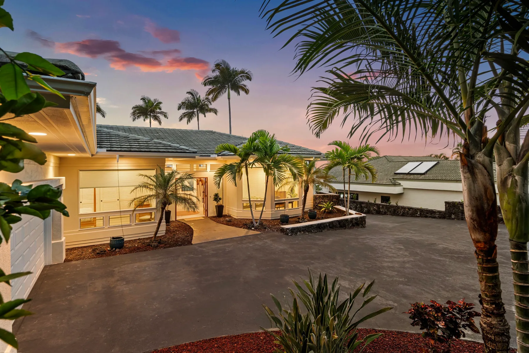 Exterior de una propiedad tropical al atardecer, con palmeras y una entrada pavimentada.