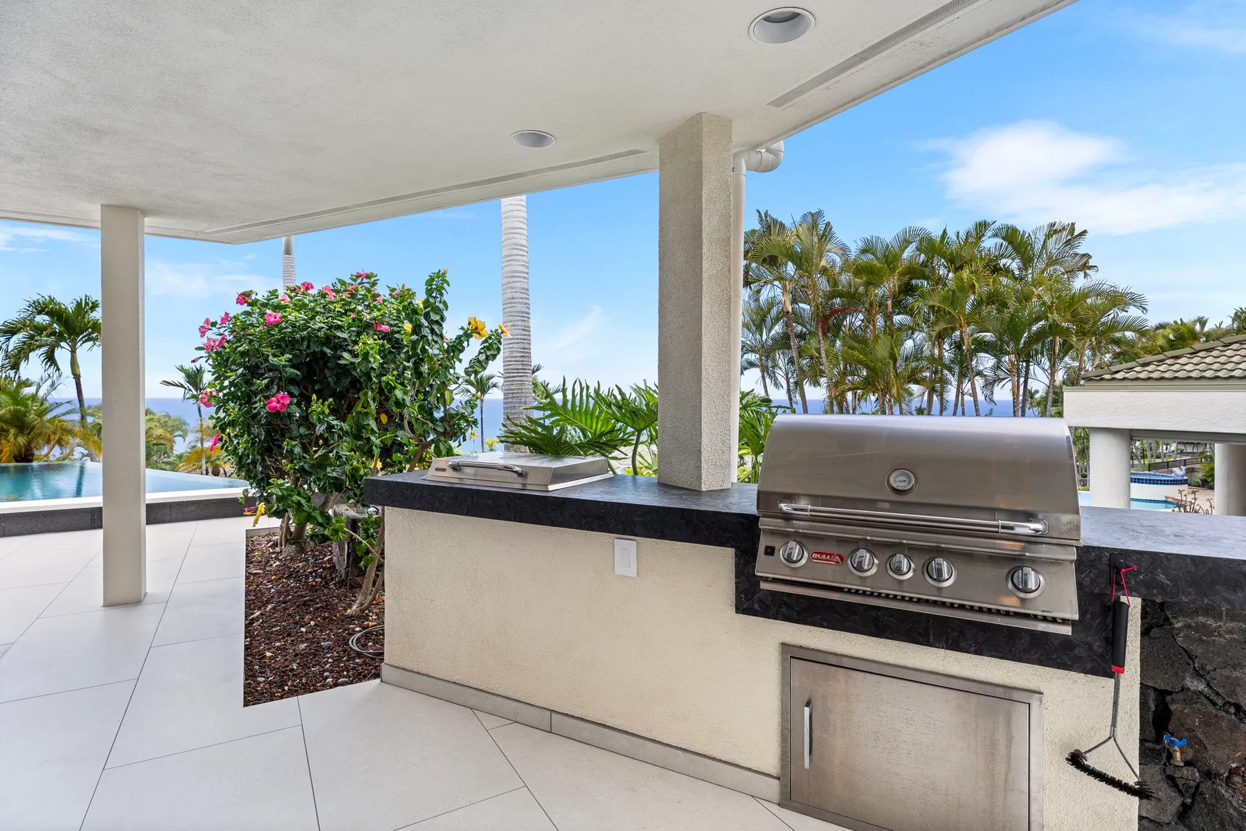 Una cocina al aire libre con parrilla empotrada da a una piscina y palmeras.