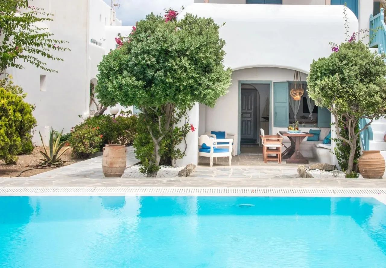 Una piscina frente a un edificio blanco con una zona de estar exterior a la sombra.