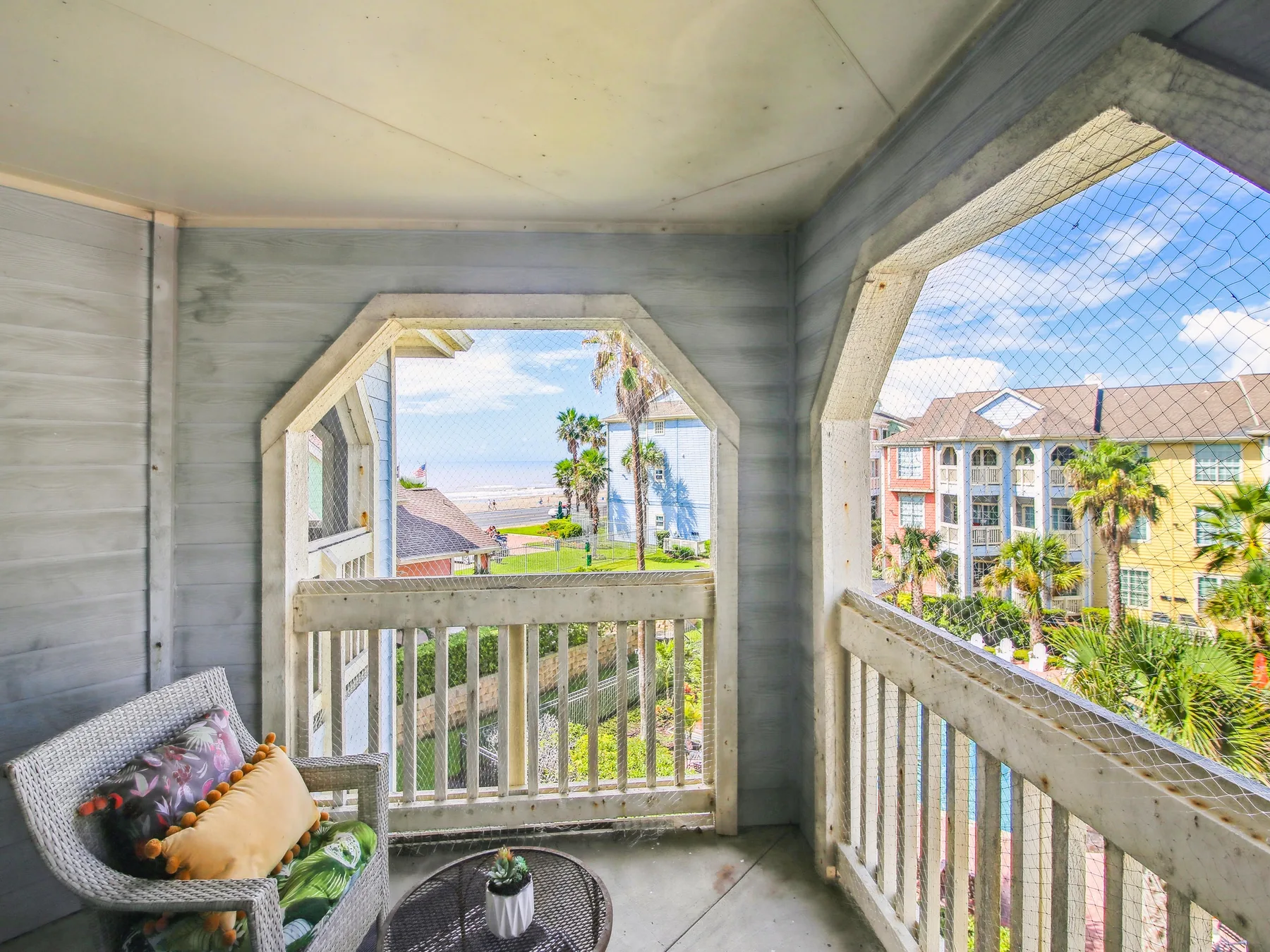 A covered balcony with seating overlooks palm trees and nearby buildings.