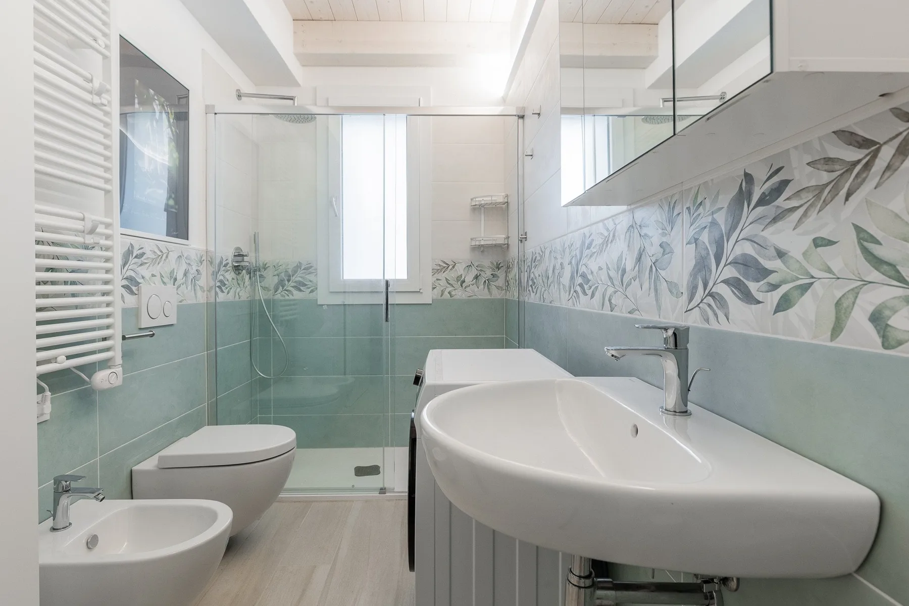 Un baño luminoso con ducha de vidrio, bañera y azulejos con hojas.