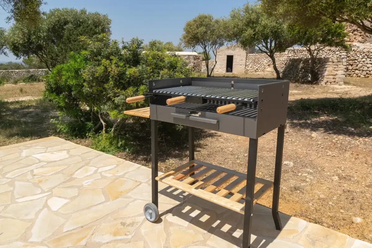 Una parrilla exterior está junto a un patio de piedra en un jardín soleado.