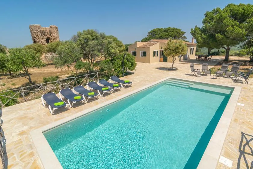 Una piscina al aire libre junto a un patio pavimentado con tumbonas y un pequeño edificio.