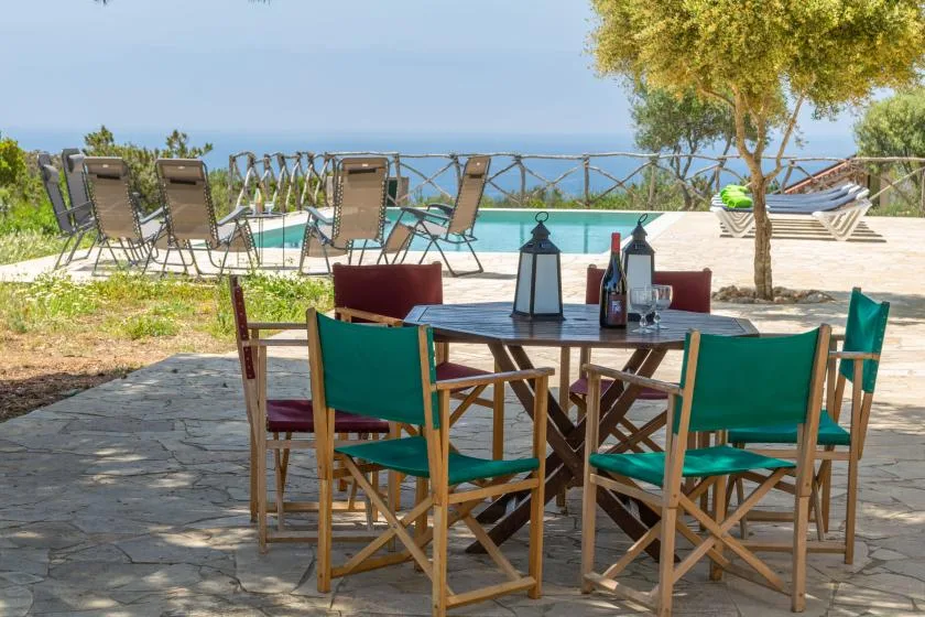 An outdoor dining area with chairs and a table overlooks a pool and the sea.