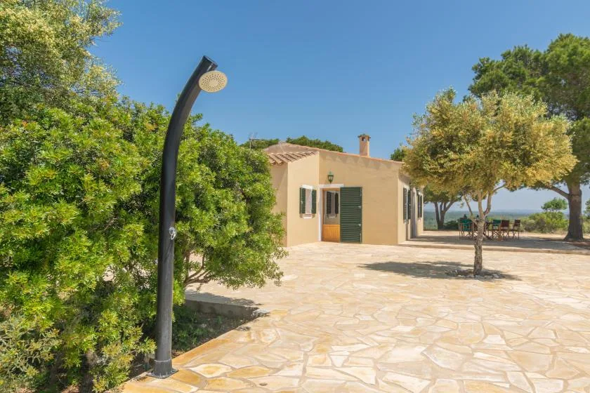 Una pequeña construcción junto a un patio empedrado, con árboles y una ducha exterior curva.