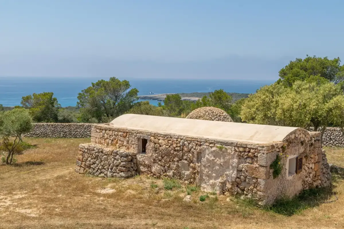 Un edificio bajo de piedra se alza en un terreno seco y abierto, con el mar a lo lejos.