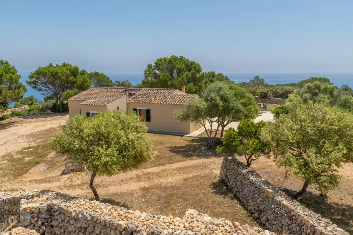 Una casa baja se encuentra entre árboles y muros de piedra en un paisaje seco.