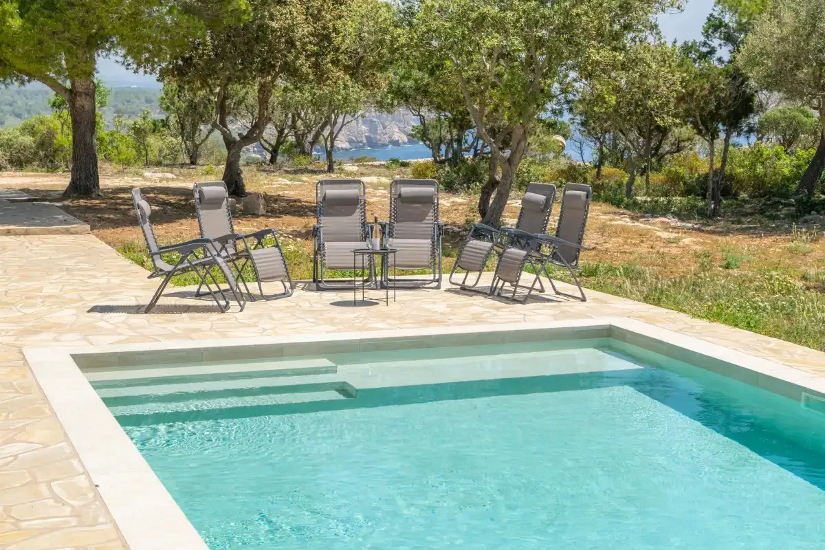 Una piscina al aire libre junto a un patio pavimentado con asientos y árboles.