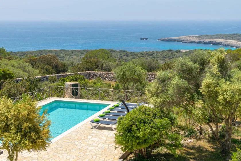 An outdoor swimming pool and loungers overlook a green coastal landscape and the sea.