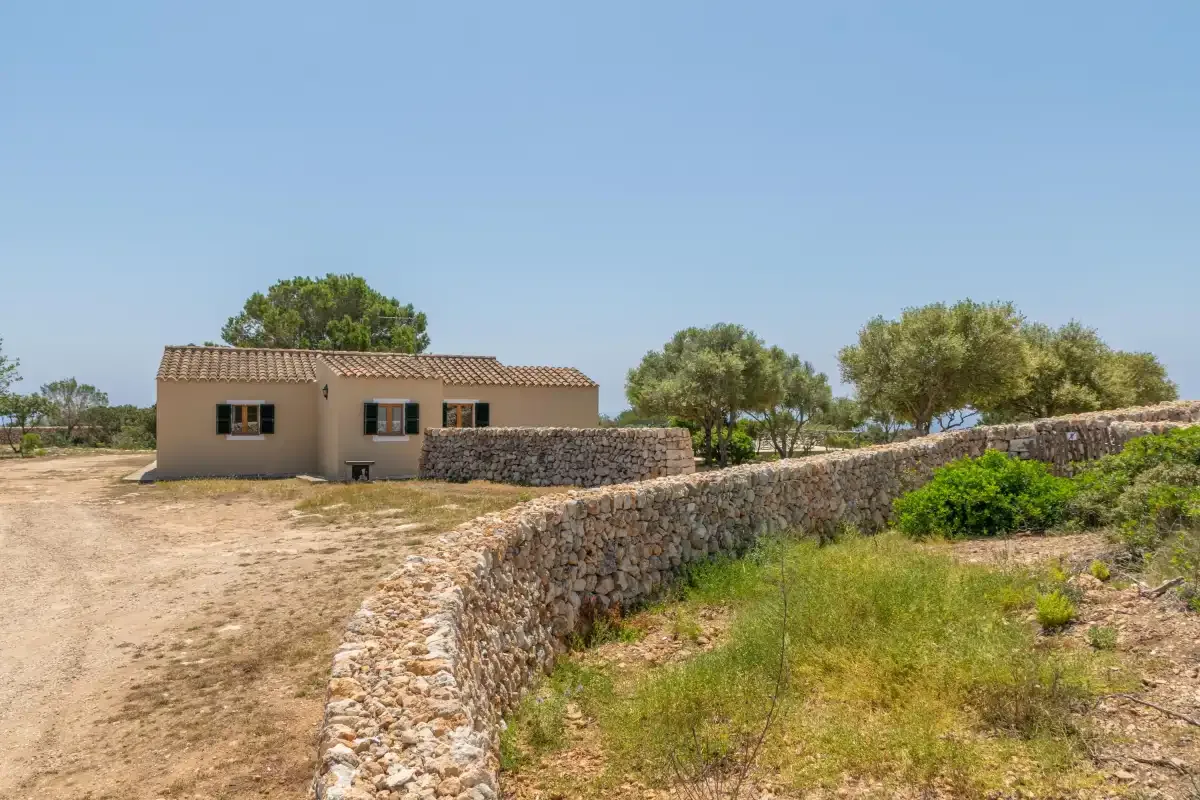 Una casa pequeña se encuentra detrás de un muro bajo de piedra en un paisaje seco y abierto.