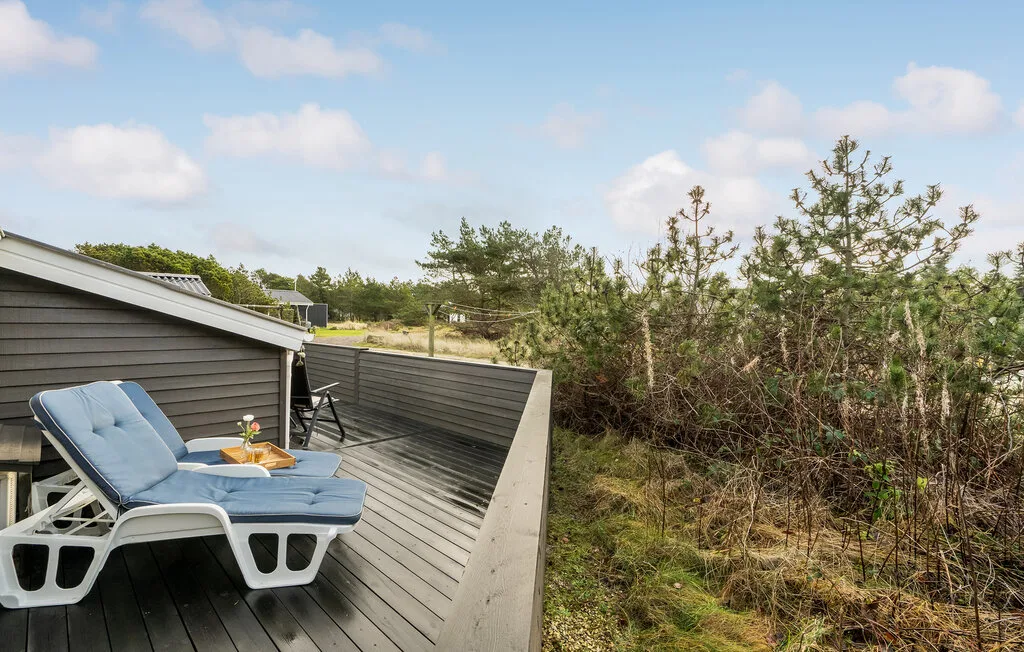 Una terraza de madera con asientos reclinables da a una zona de arbustos bajo un cielo azul.