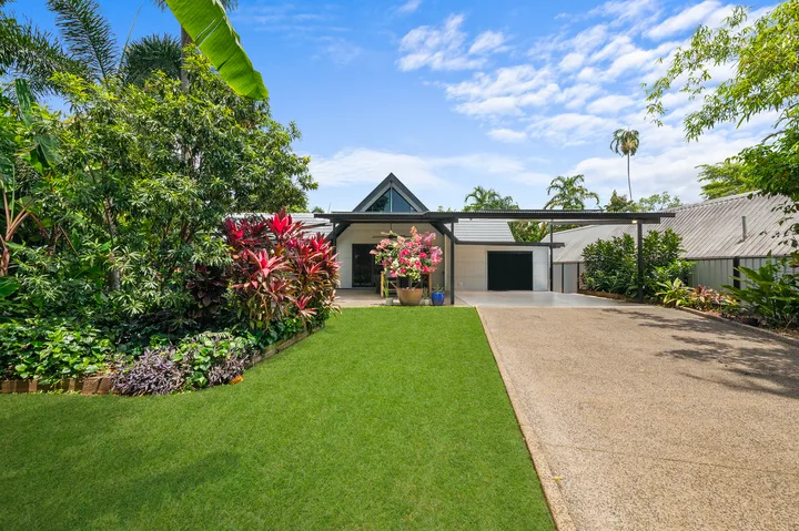 Una casa moderna se encuentra detrás de un césped verde y un jardín tropical ajardinado.
