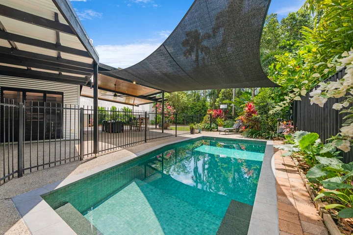 Una piscina exterior está junto a un jardín con plantas y bajo una cubierta de sombra.