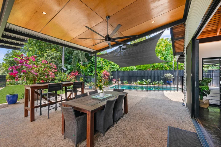 Una zona cubierta de comedor al aire libre da a una piscina y un jardín con plantas.