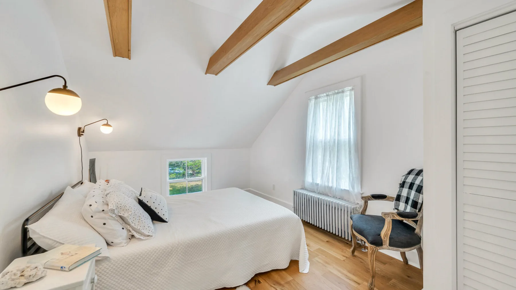 Un dormitorio luminoso con una cama bajo vigas de madera y una ventana con cortinas.