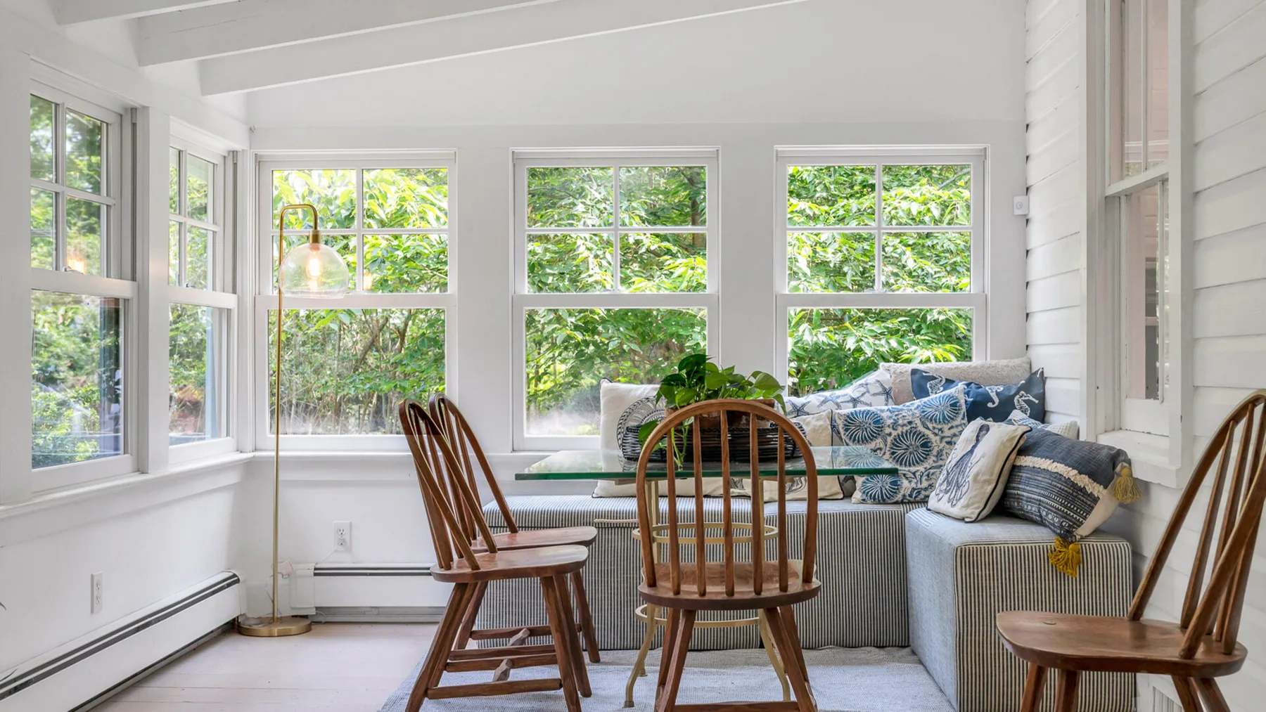 A bright sunroom has large windows, a seating area, and wooden chairs.