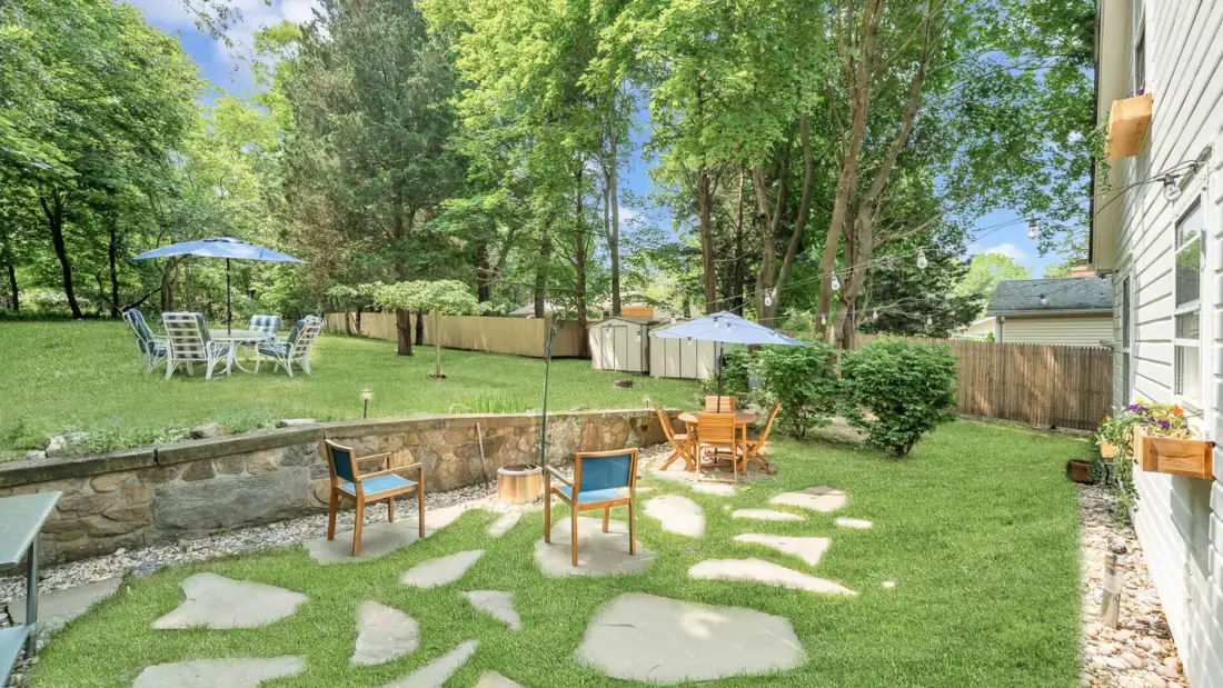 Un patio trasero con césped tiene senderos de piedra, asientos al aire libre y árboles.