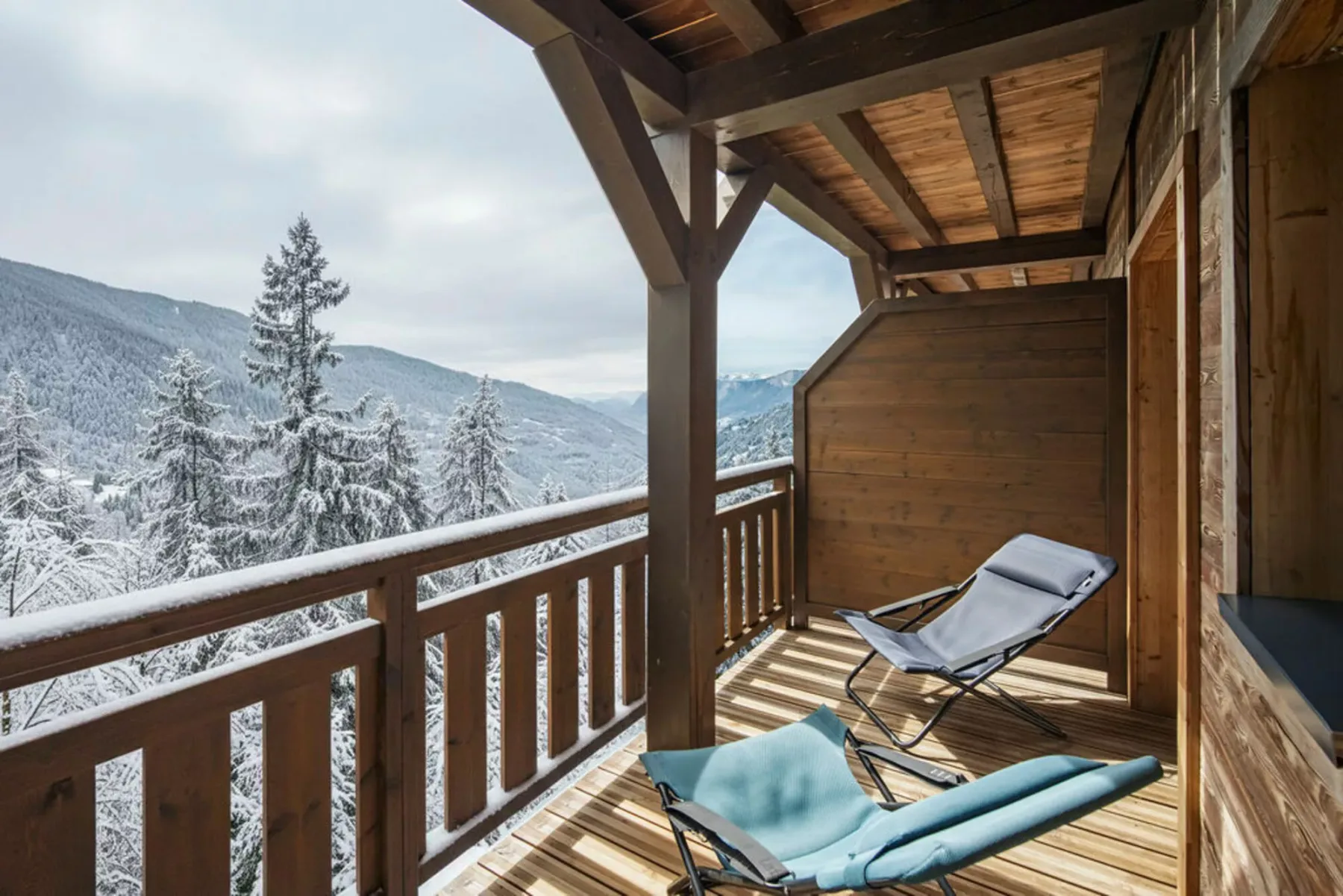 A covered wooden balcony overlooks snow-covered trees and mountain scenery.