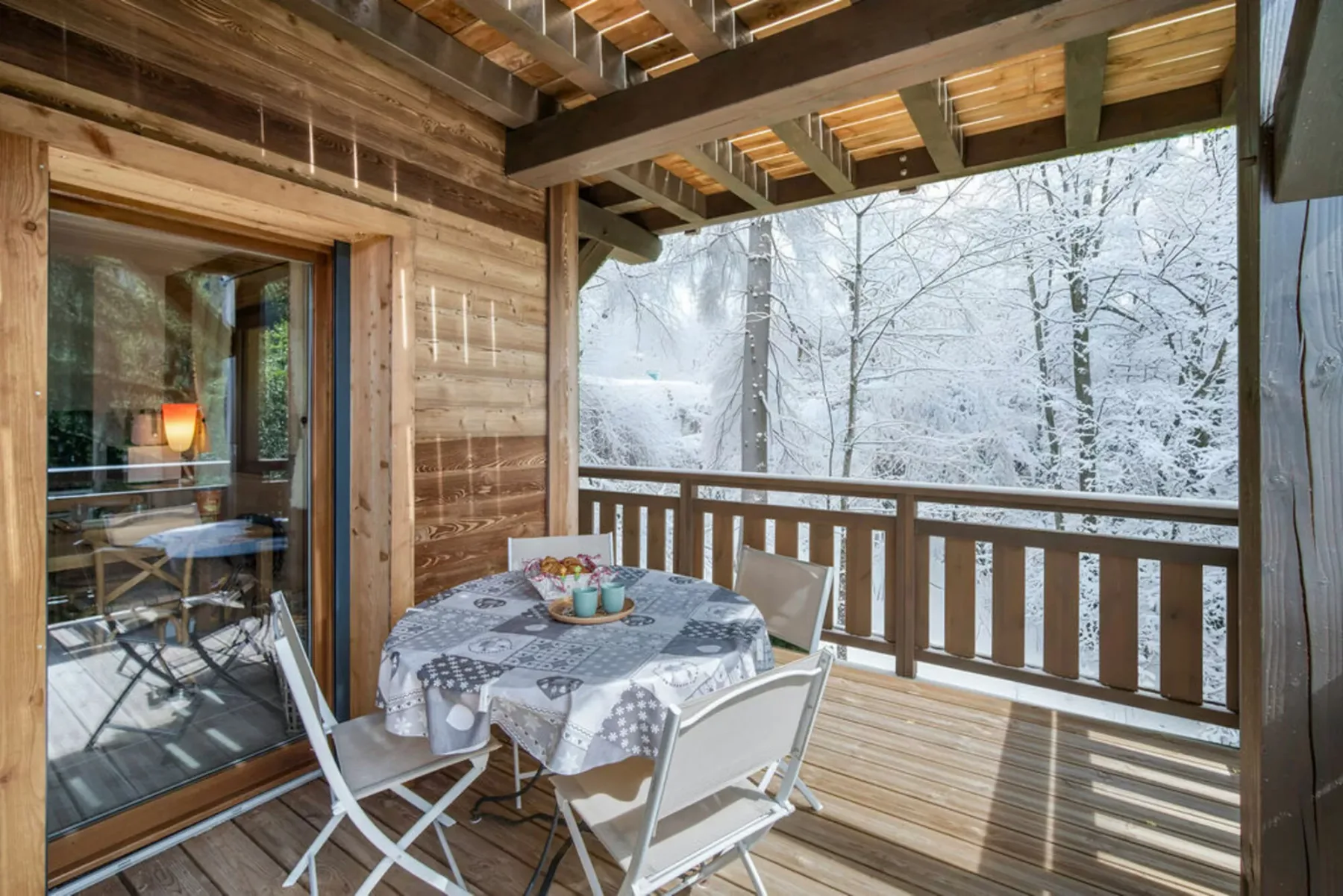 A covered wooden deck has a dining table and snowy trees beyond the railing.