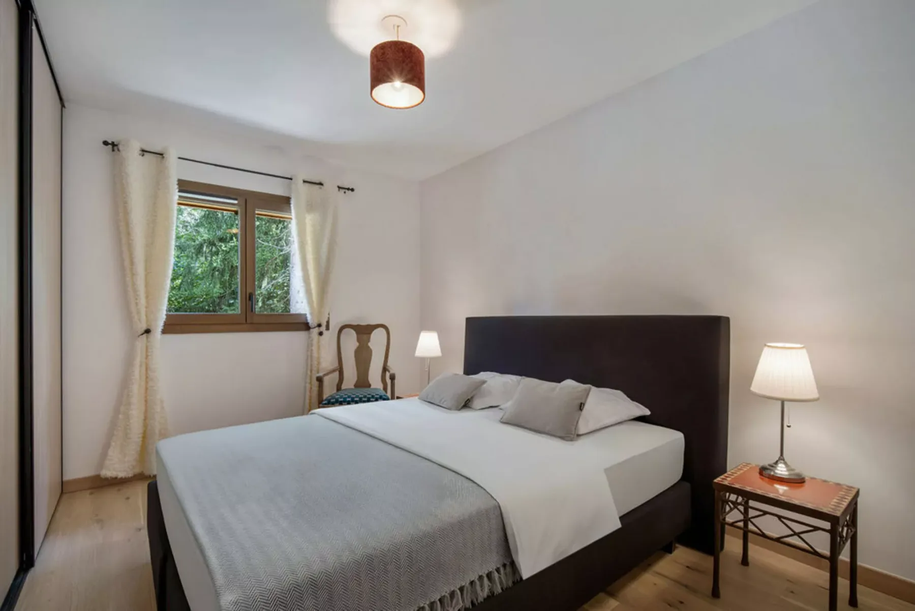 A bedroom with a neatly made bed beside a window overlooking greenery.