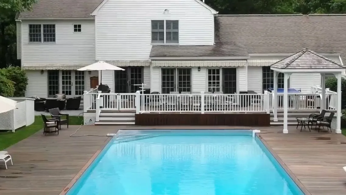 Una piscina está junto a una terraza de madera, frente a una casa blanca.