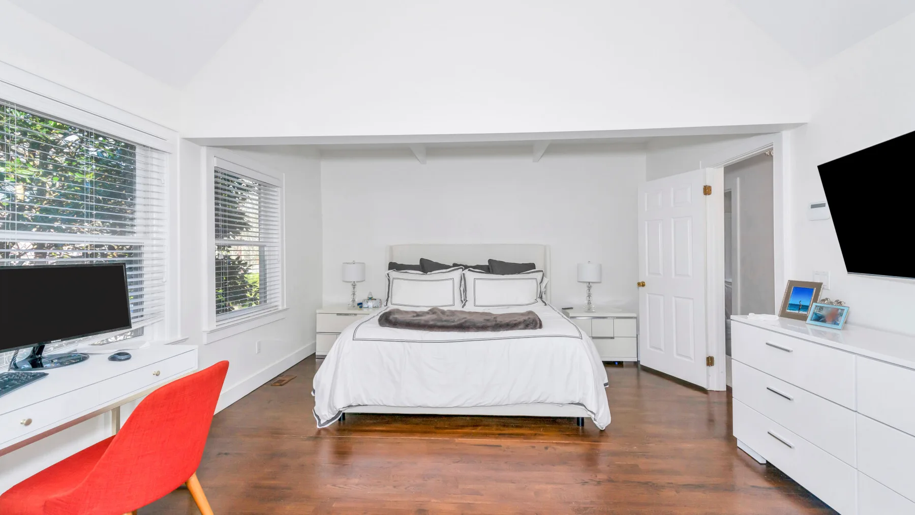 Un dormitorio luminoso con una cama tendida, muebles blancos y una silla roja.