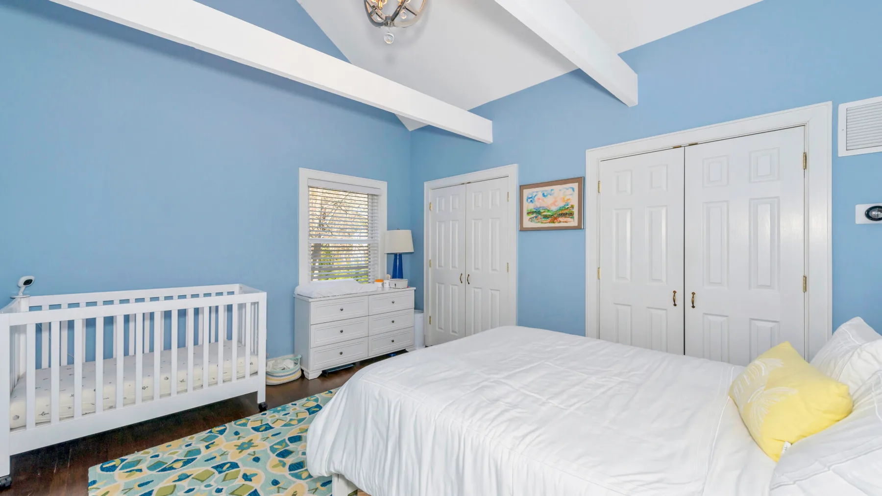 Un dormitorio luminoso con una cama blanca, una cuna y paredes azules.
