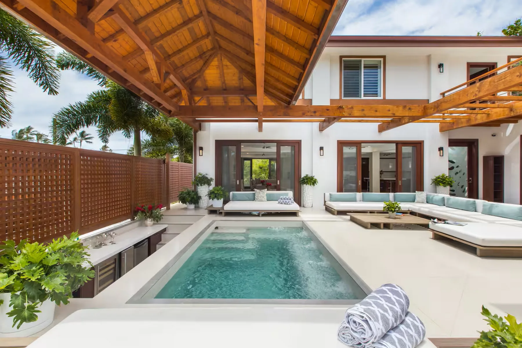 A courtyard pool sits beside a white house beneath a wooden pergola.