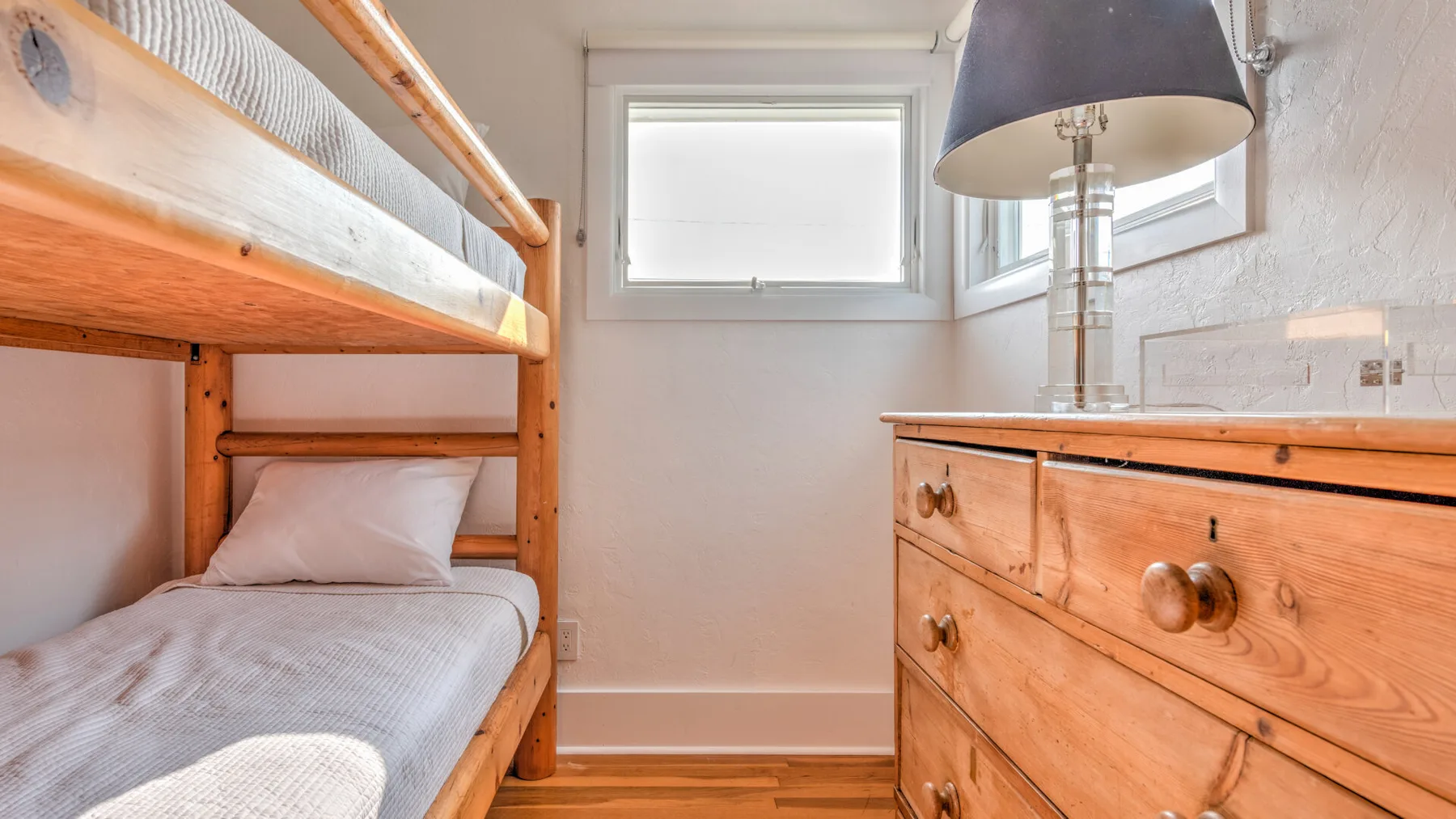 Un dormitorio luminoso con una litera de madera, una cómoda y ventanas.