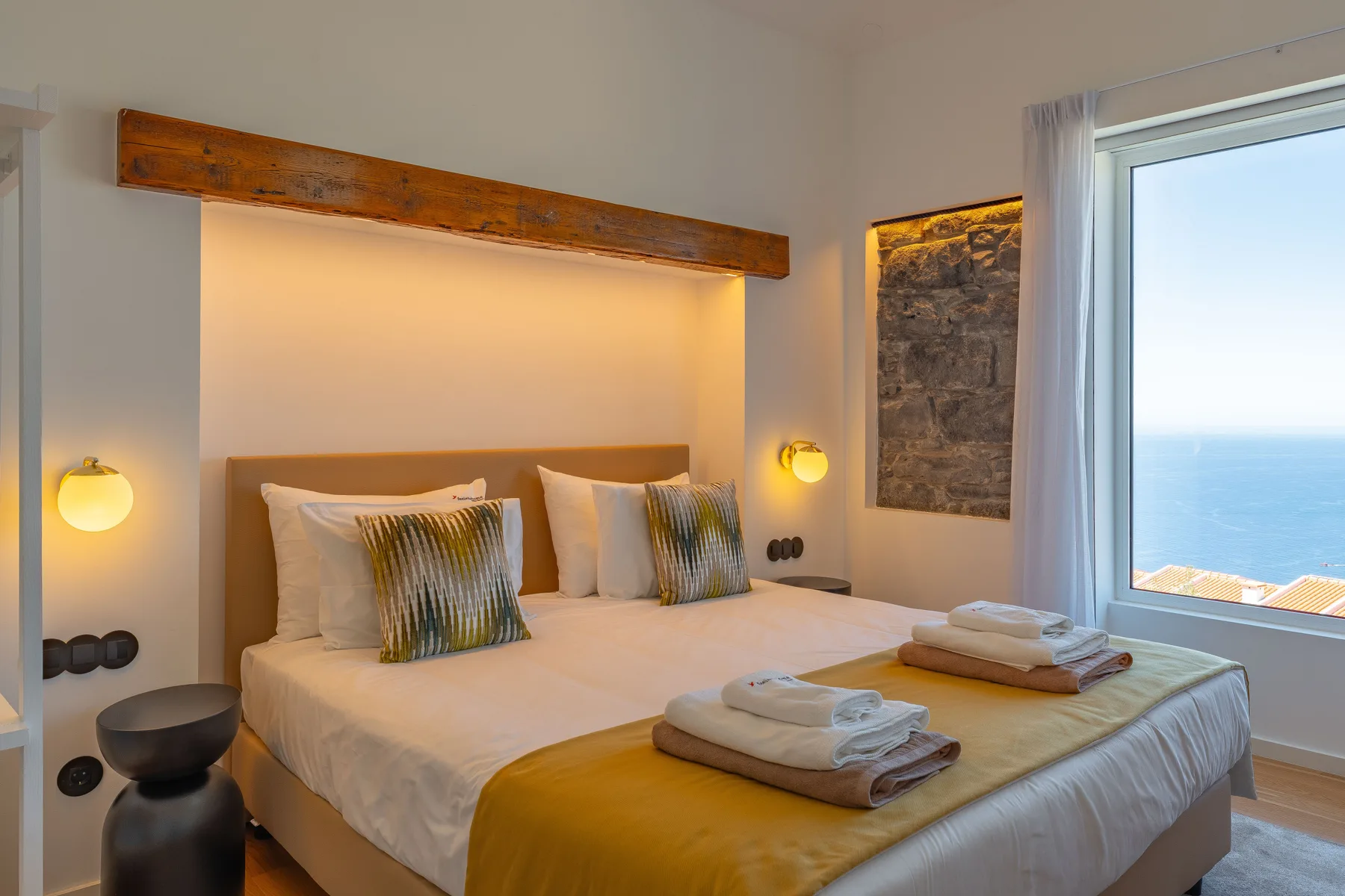 A bedroom with a neatly made bed and a large window overlooking the water.