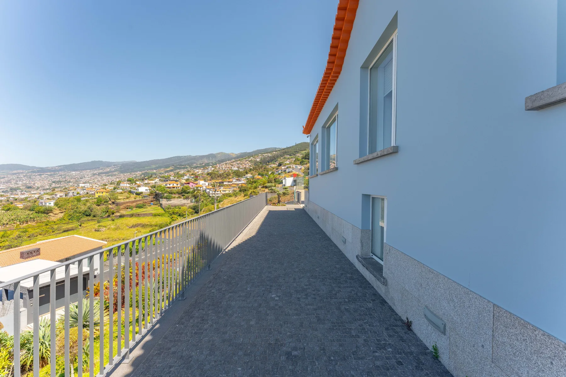 A paved balcony runs alongside a white building, overlooking a hillside neighborhood.